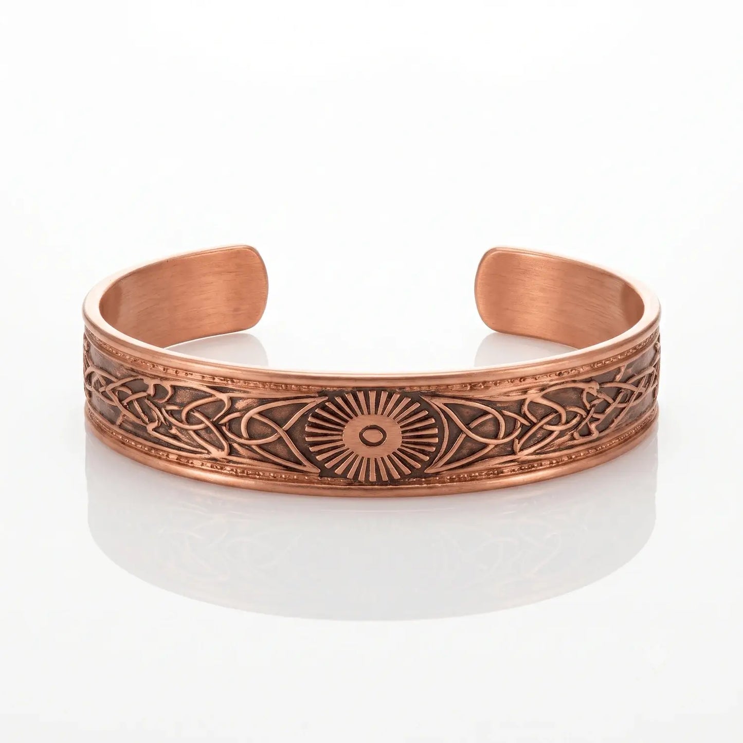 A copper-toned open cuff bracelet with engraved Celtic knotwork patterns and a central sunburst motif, displayed on a clean white reflective surface.